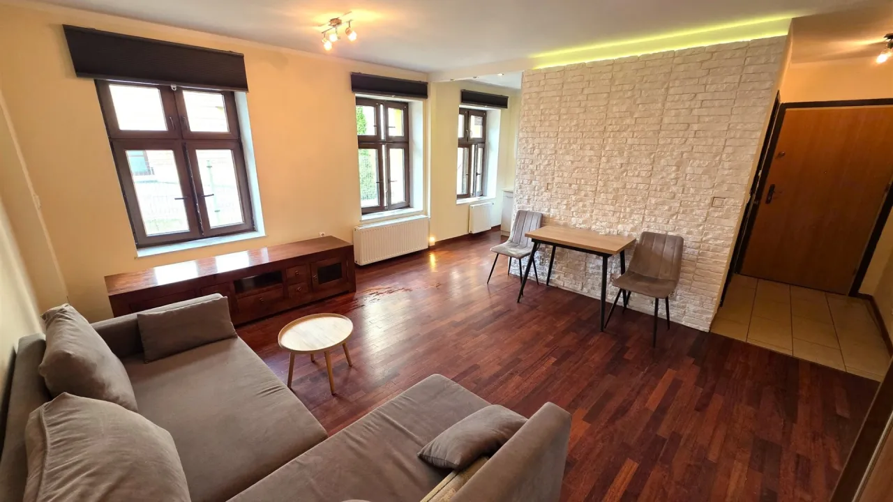 Nowe mieszkanie na wynajem: salon z kanapą, stolikiem, biurkiem i krzesłami. Widok na okna i drzwi wejściowe.