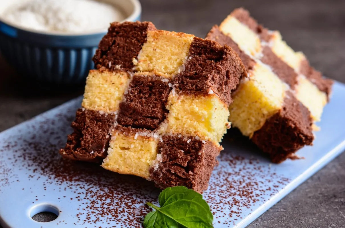 Ciasto szachownica z bitą śmietaną przepis. Dwa kawałki ciasta w kratkę, czekoladowo-waniliowe, na niebieskiej desce, posypane kakao.