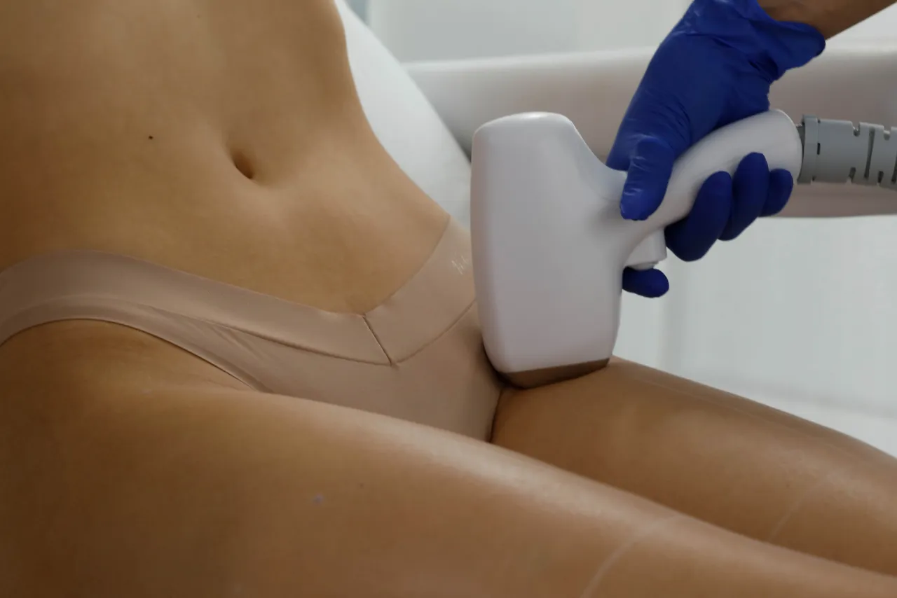 Zabieg depilacji laserowej okolic bikini. Dłoń w rękawiczce trzyma depilator, który zbliża się do skóry. To może być odpowiedź na pytanie, jaka depilacja bikini najlepsza.