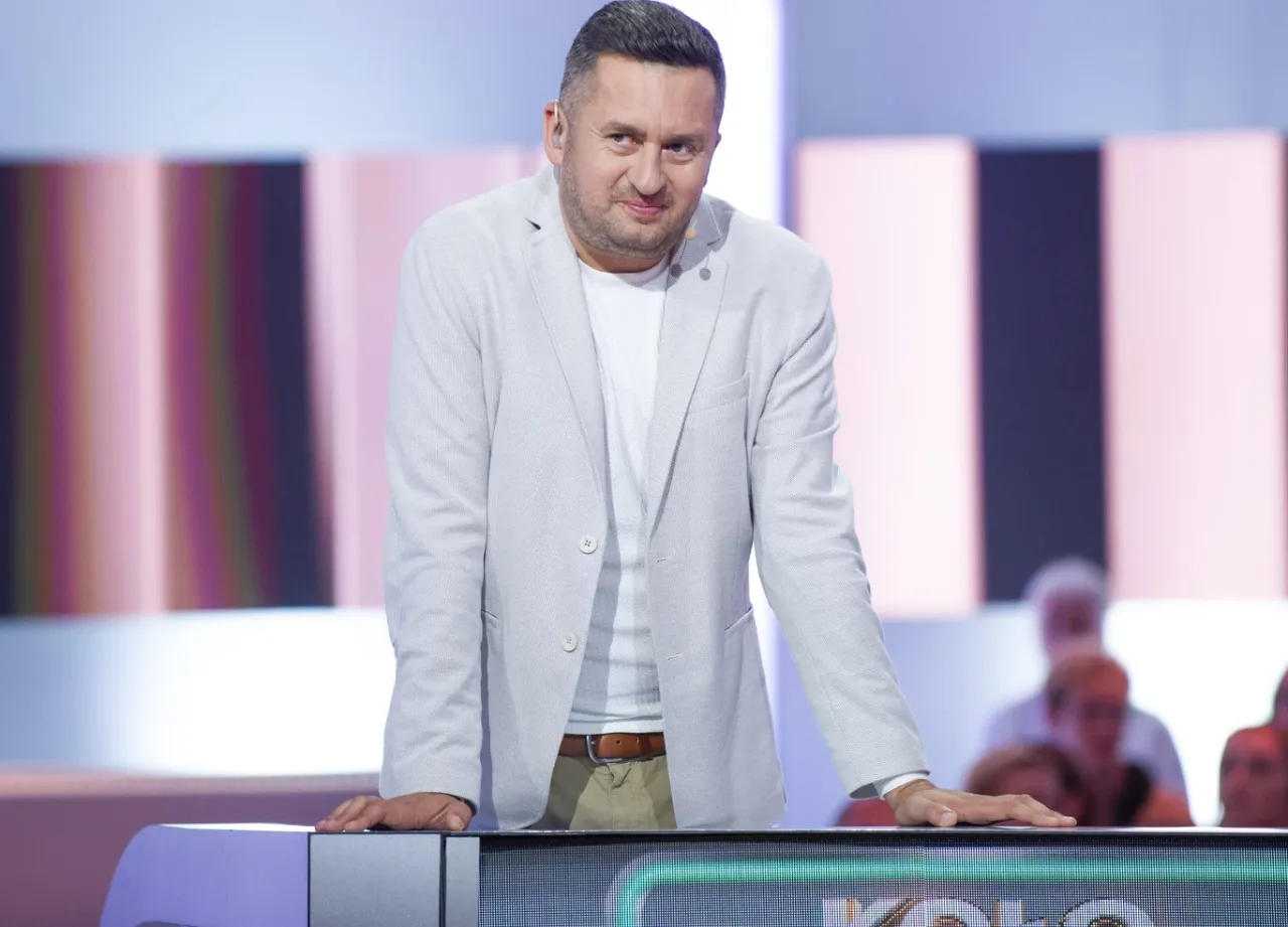 Mężczyzna w jasnej marynarce, który prowadzi Koło Fortuny, z uśmiechem spogląda na widownię.