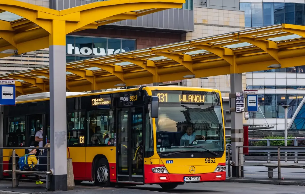 Ż&oacute;łto-czerwony autobus miejski na przystanku. Kierowca autobusu, jak i pasażerowie, zastanawiają się, ile zarabia kierowca autobusu.