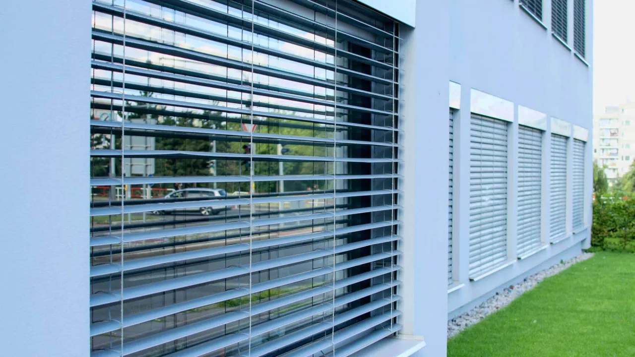 Nowoczesne żaluzje fasadowe zewnętrzne chronią budynek przed słońcem. W odbiciu widać ulicę i samochód.