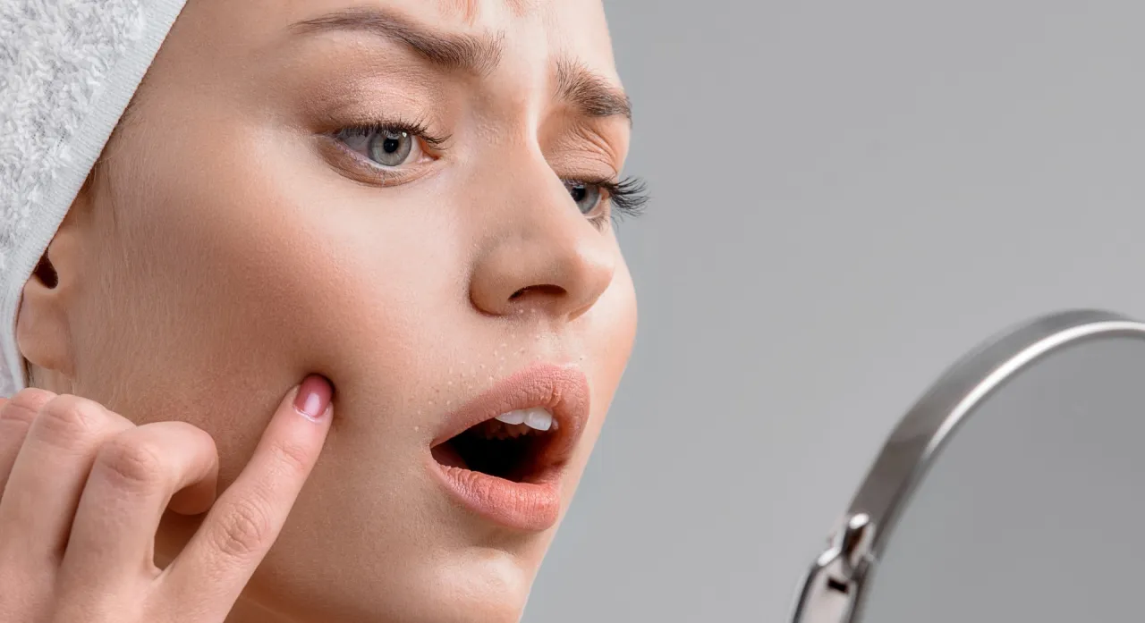 Zastanawiasz się, jaki wosk do depilacji twarzy będzie najlepszy? Kobieta z ręcznikiem na głowie ogląda w lustrze niedoskonałości sk&oacute;ry.