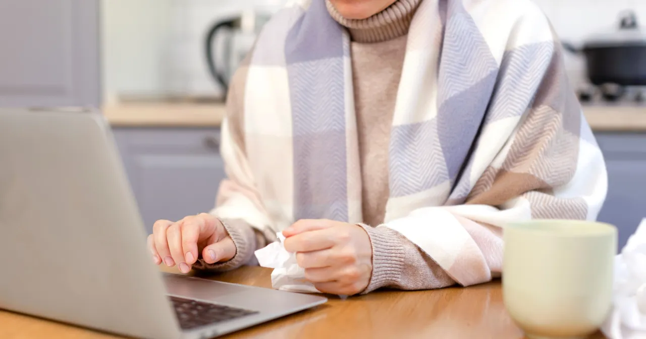 Osoba owinięta kocem siedzi przy laptopie, trzymając chusteczkę. Zastanawia się, czy l4 w weekend się opłaca.