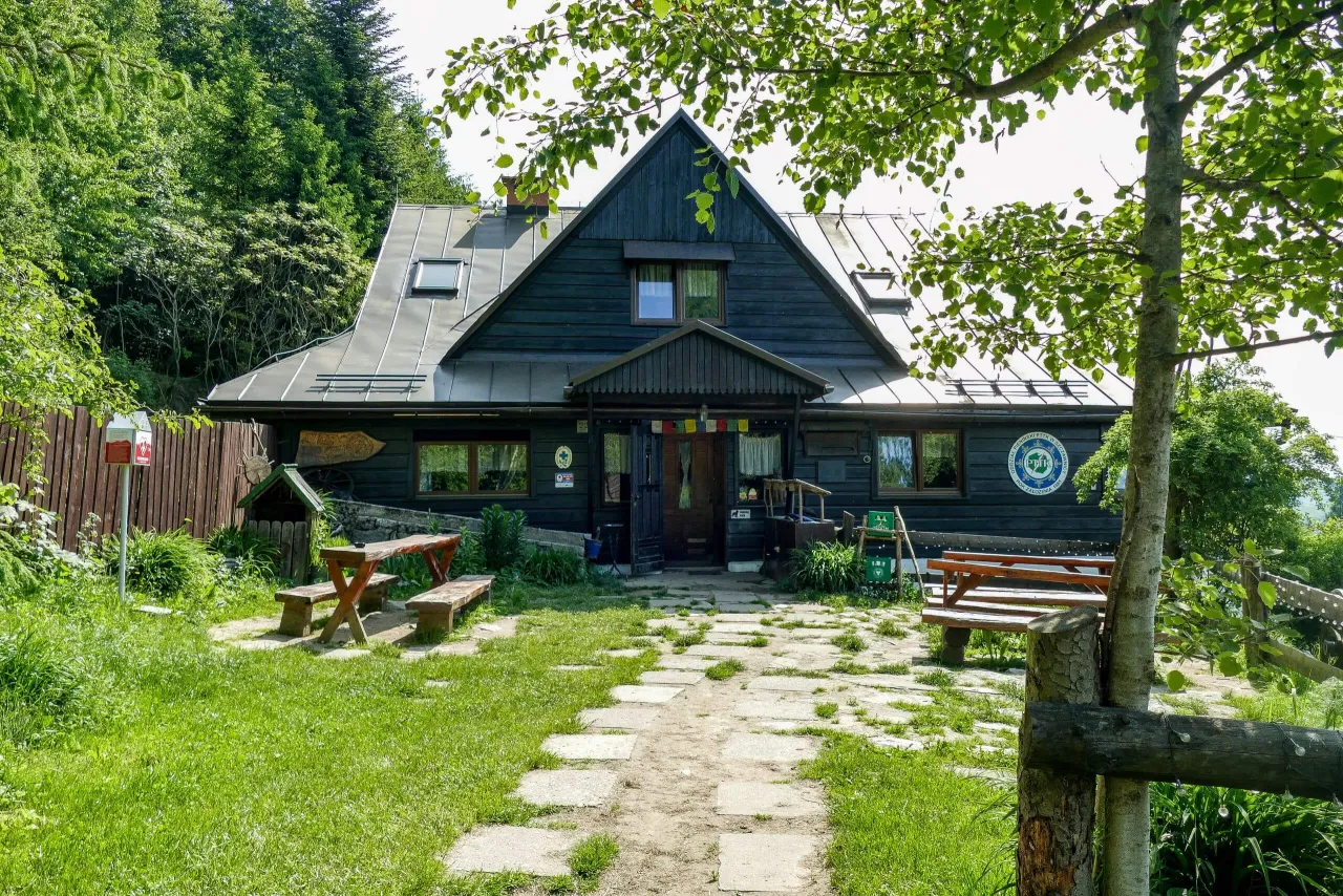 Drewniana bac&oacute;wka pod Bereśnikiem otoczona zielenią. Przed budynkiem kamienna ścieżka, ławki i stoły.