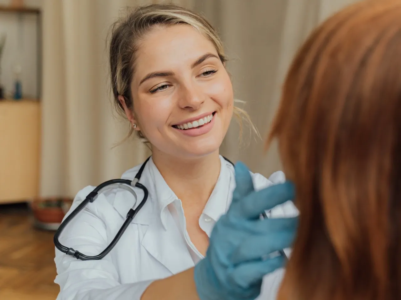 Uśmiechnięta lekarka w białym kitlu i niebieskich rękawiczkach bada pacjenta. Czy 16 latek może sam iść do lekarza? Tak, to normalne.
