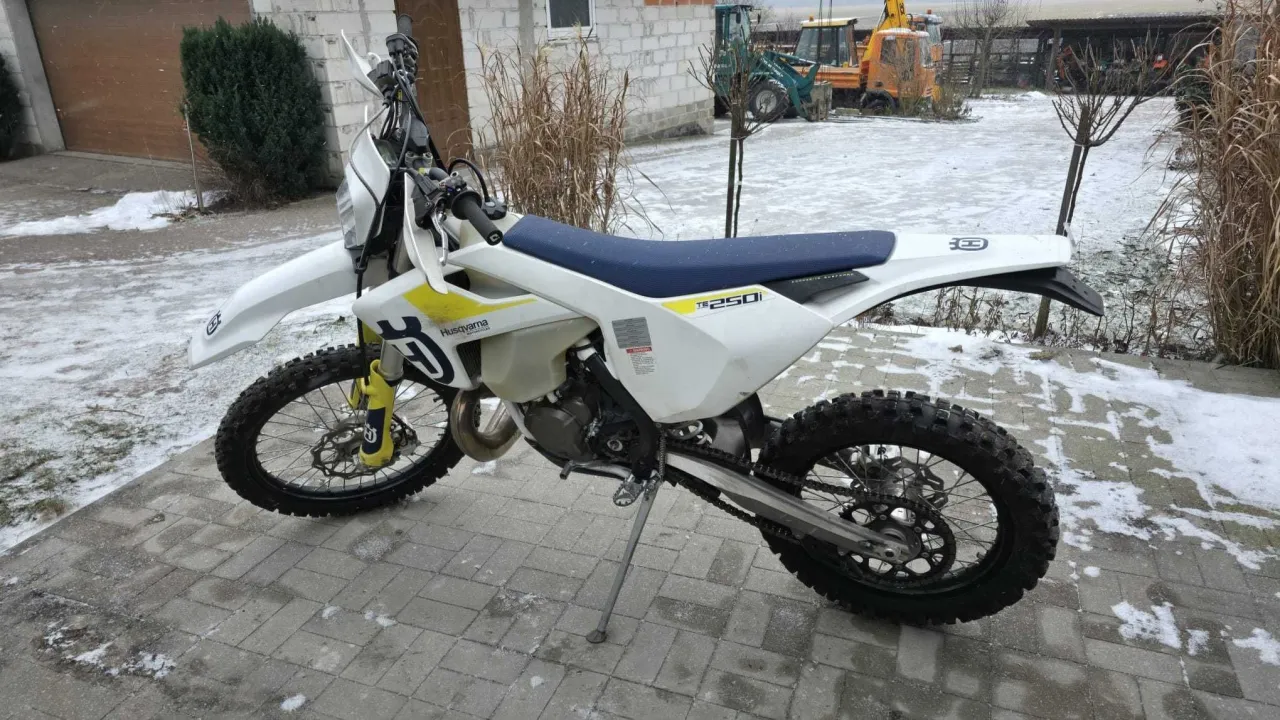 Husqvarna 250 cross gotowy do jazdy po śniegu. Biały motocykl z niebieskim siedzeniem stoi na kostce brukowej, w tle widać maszyny budowlane.