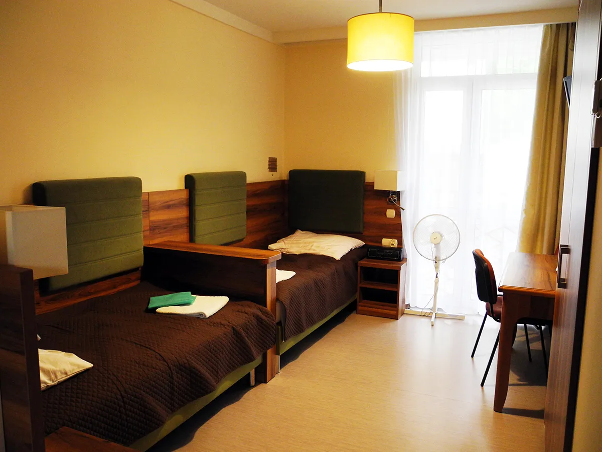 Pok&oacute;j w sanatorium Rafał Busko Zdr&oacute;j z dwoma ł&oacute;żkami, biurkiem i wentylatorem.