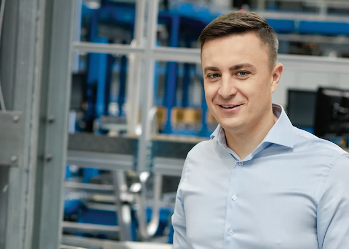 Uśmiechnięty mężczyzna w jasnoniebieskiej koszuli, na tle hali produkcyjnej. To pracownik firmy X KOM, kt&oacute;ra zajmuje się technologiami.