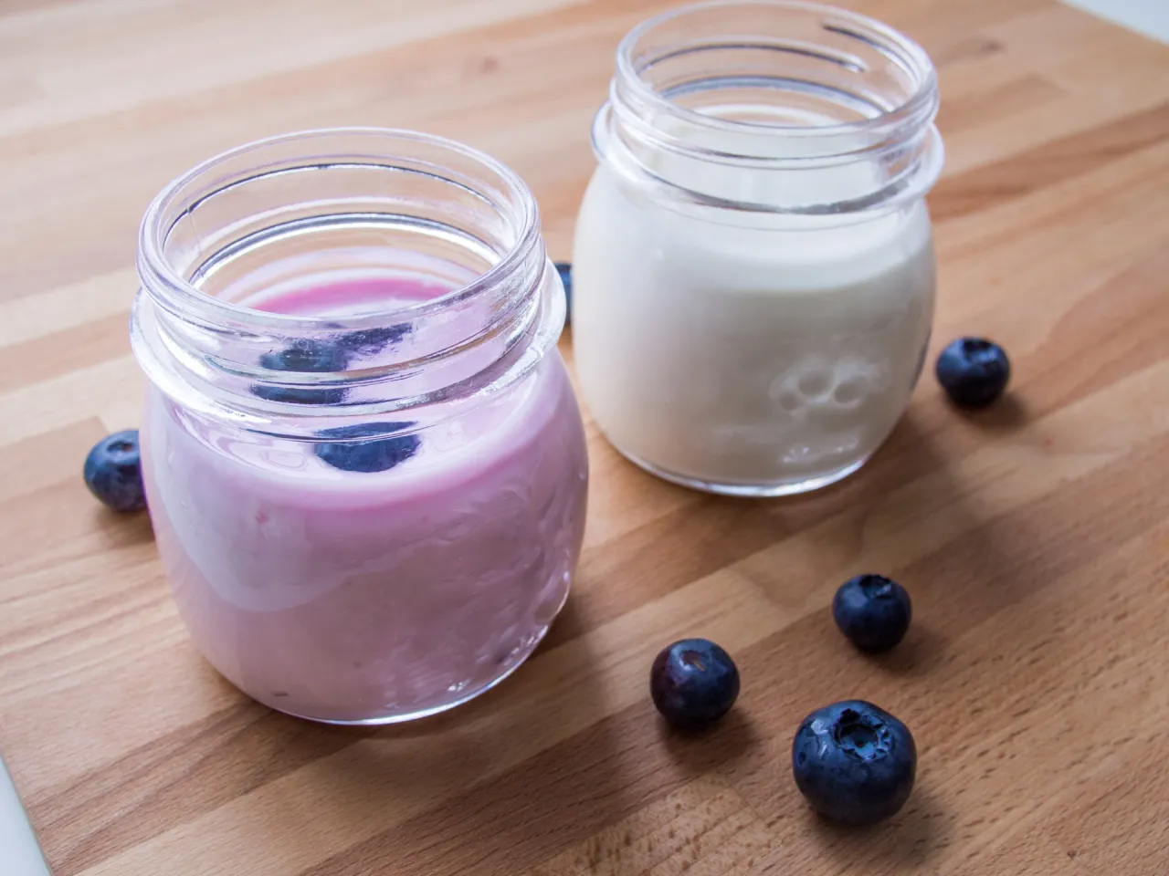 Dwa słoiczki z jogurtem, jeden r&oacute;żowy z jagodami, drugi biały. Wok&oacute;ł świeże bor&oacute;wki. Wegańskie kultury bakterii jogurtowych co to jest?