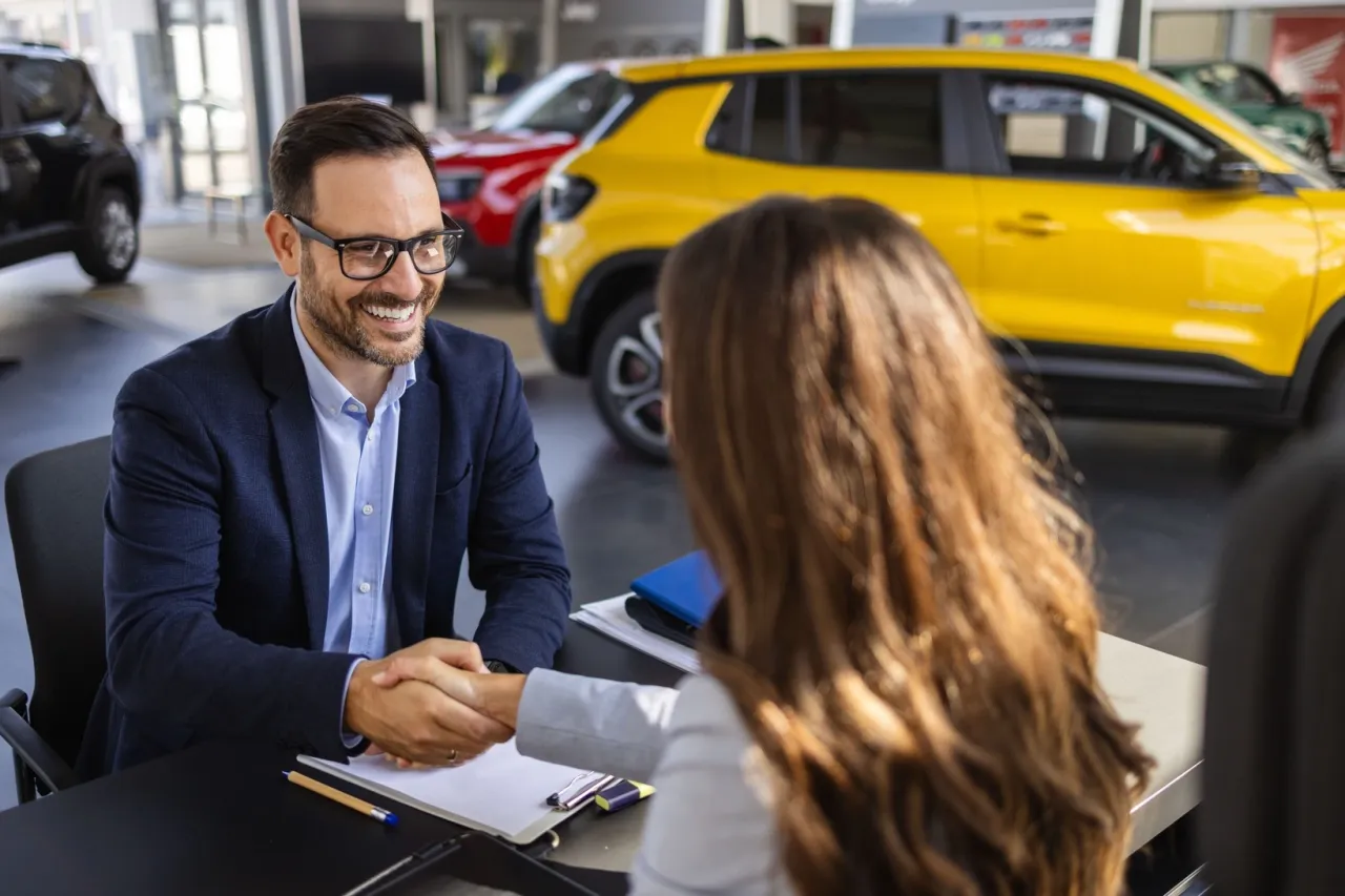 Uśmiechnięty sprzedawca samochod&oacute;w ściska dłoń klientki, omawiając leasing bez wpłaty własnej. W tle ż&oacute;łty samoch&oacute;d.