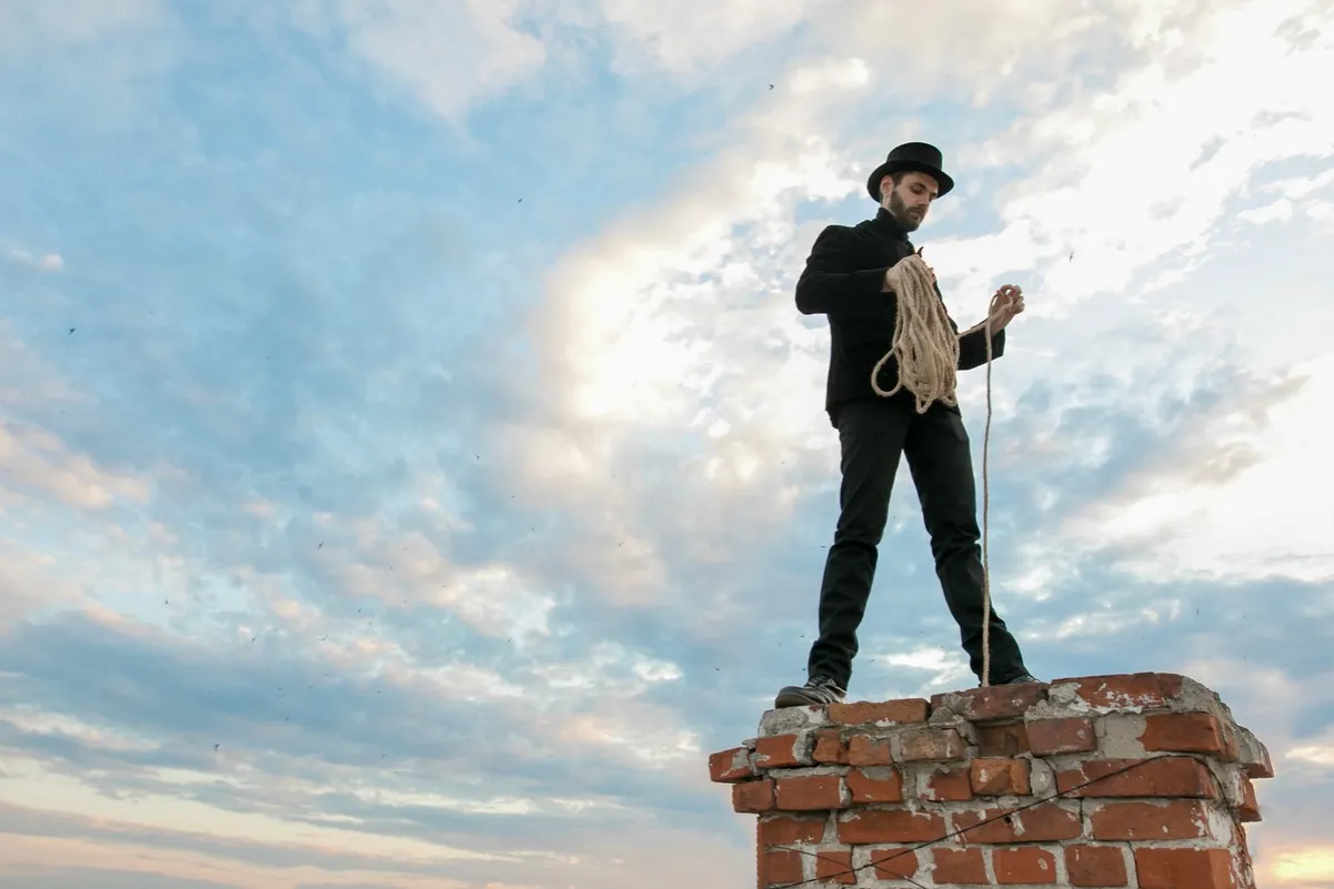 Mężczyzna w cylindrze i czarnym garniturze stoi na szczycie ceglanego komina, trzymając linę. To, czego nie czyści kominiarz, to chmury i ptaki na niebie.