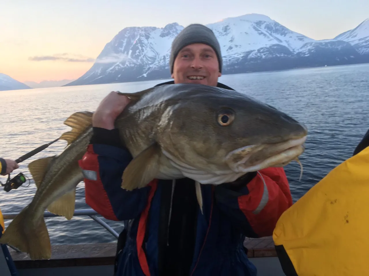 Szczęśliwy wędkarz z wielkim dorszem na tle norweskich gór. Pomyśl, co zabrać na ryby do Norwegii, by złowić takie okazy!