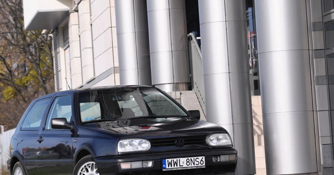 Czarny Volkswagen Golf III, jeden z pierwszych aut tej generacji, zaparkowany przed nowoczesnym budynkiem z metalowymi kolumnami.