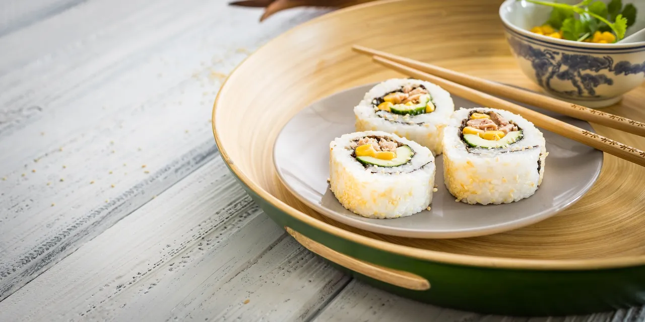 Trzy rolki sushi california roll z sezamem, ogórkiem i tuńczykiem na szarym talerzyku, obok pałeczki i miseczka z zieleniną.