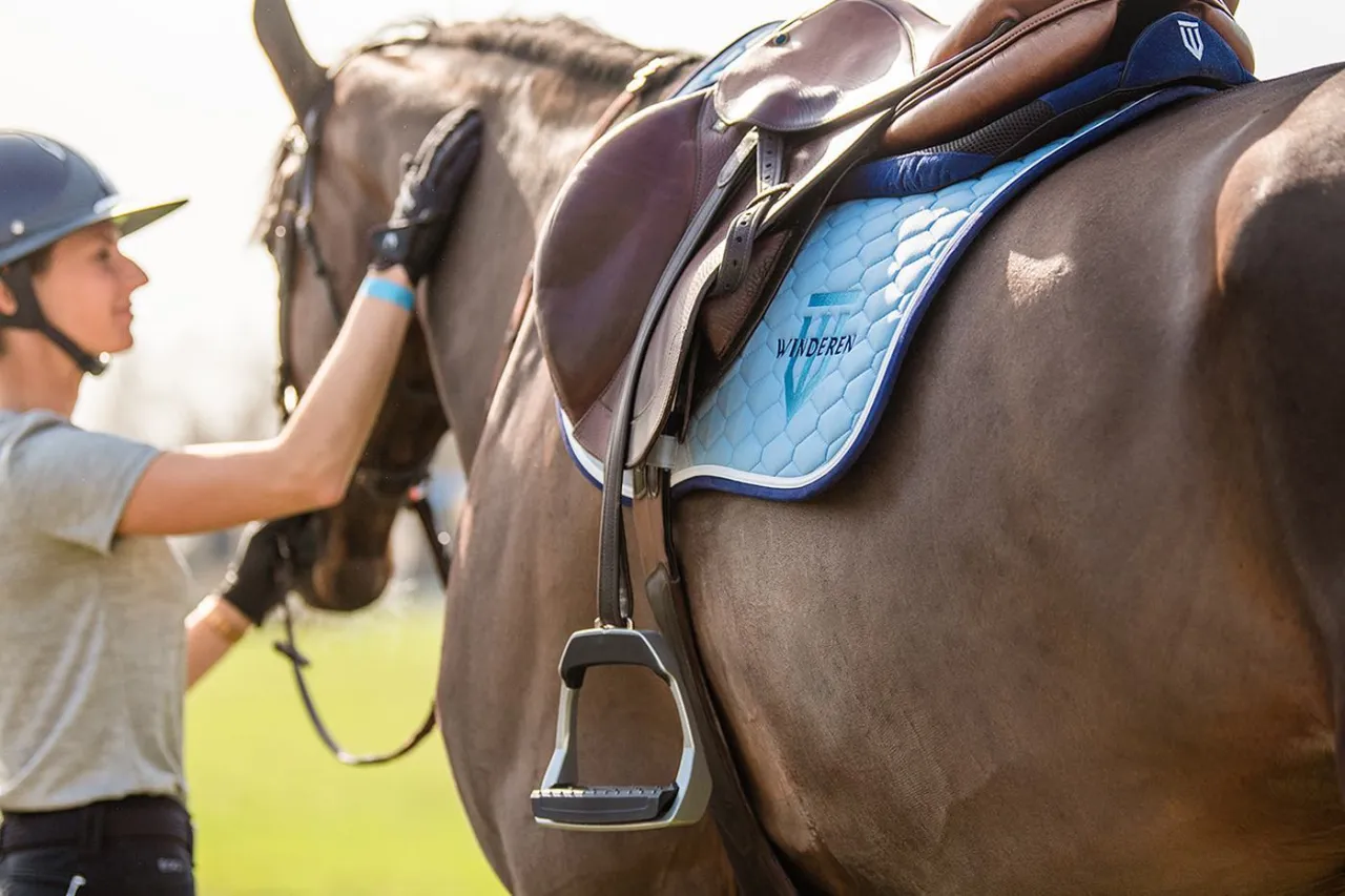 Jeździec głaszcze konia. Na grzbiecie zwierzęcia widać siodło i jasnoniebieski czaprak z logo.