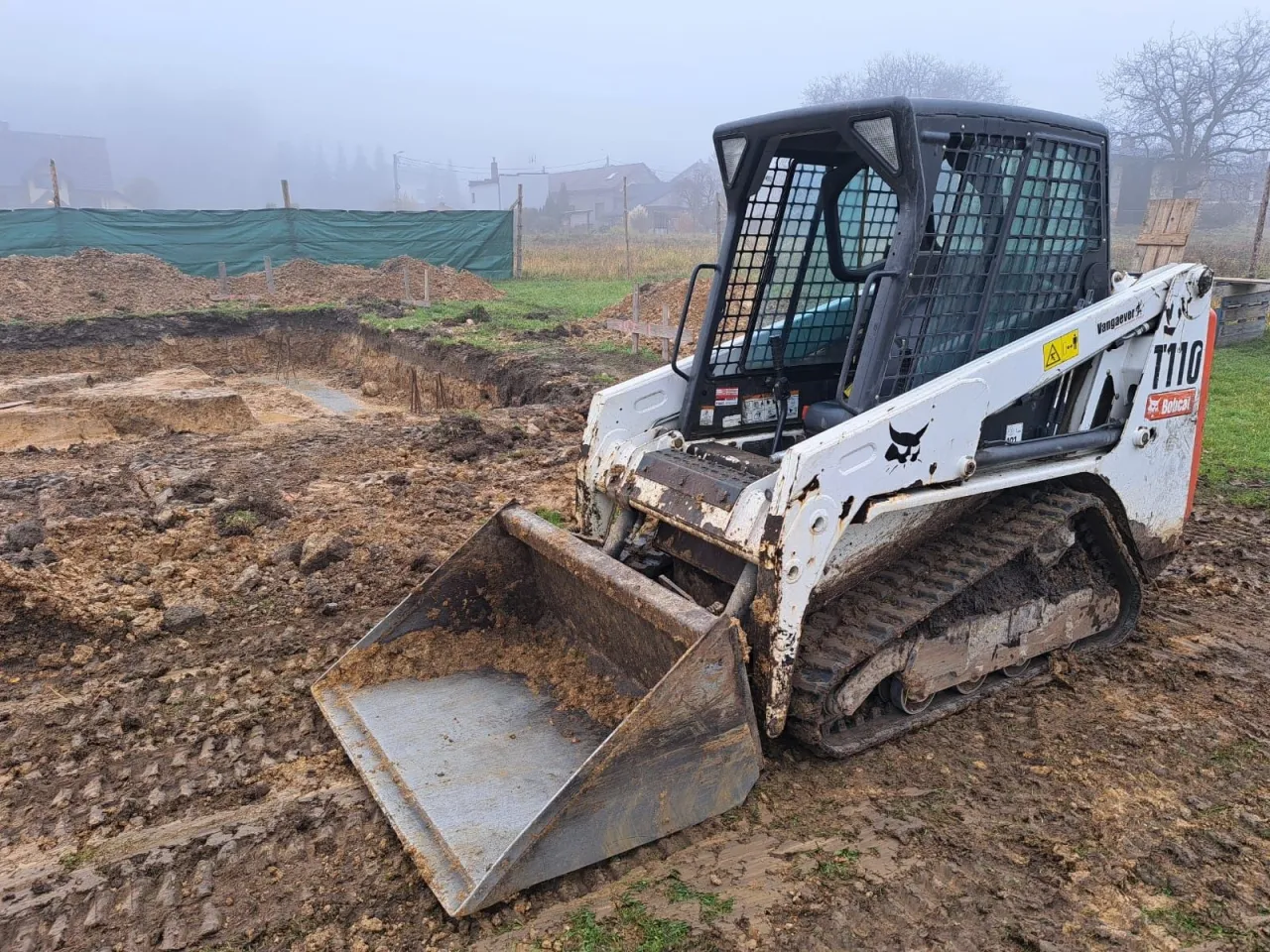Biała minikoparka Bobcat T110 na gąsienicach stoi na błotnistym placu budowy. W tle wykop pod fundamenty i zielona siatka.