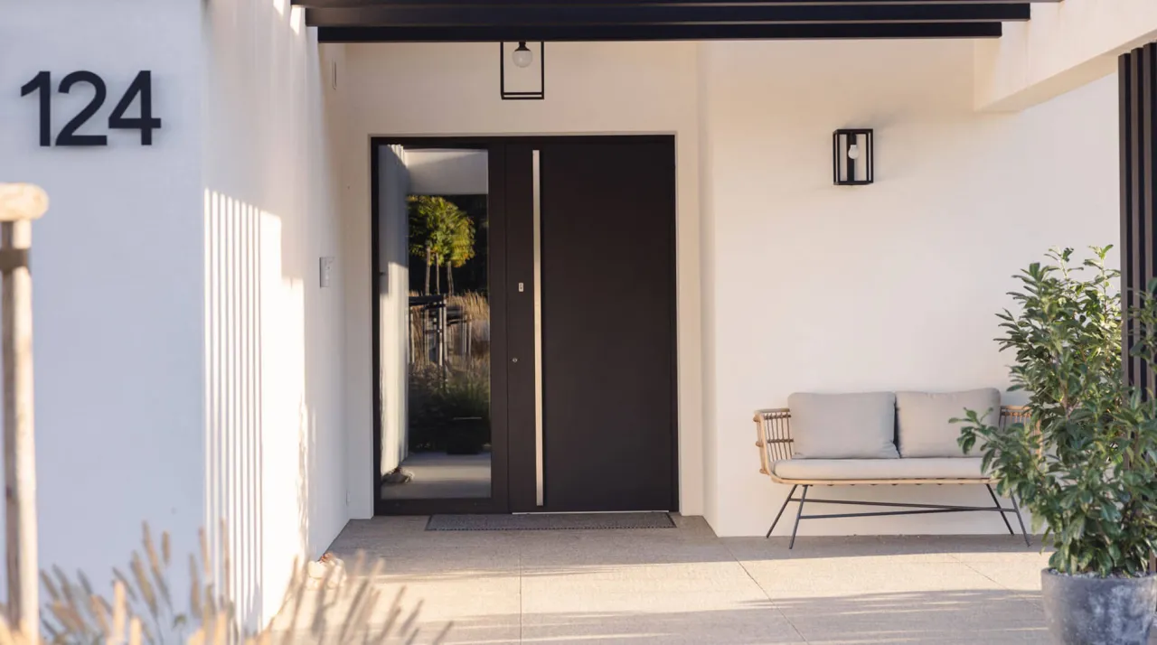 Nowoczesne drzwi zewnętrzne w kolorze grafitowym, idealnie wpisujące się w najmodniejsze kolory drzwi zewnętrznych. Obok ławka i roślinność.