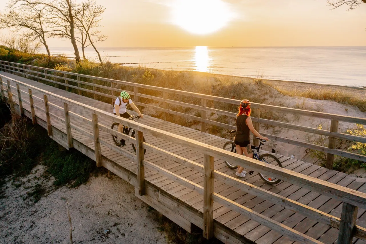 Dwie osoby na rowerach przemierzają drewnianą trasę w Sarbinowie o zachodzie słońca. Piękny widok na morze.