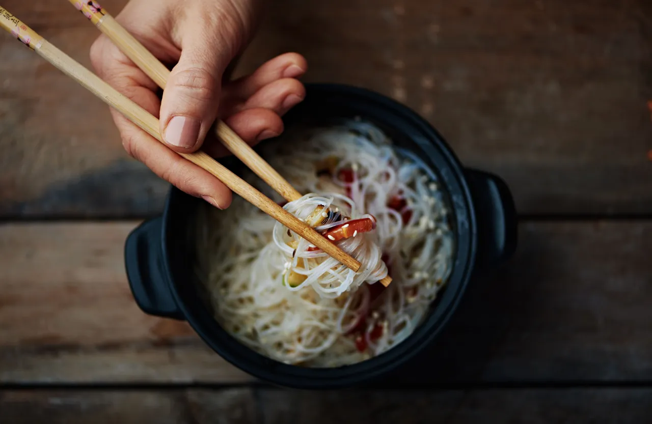 Dłoń trzyma pałeczki nad miską z makaronem ryżowym. Czy makaron sojowy jest zdrowy? To danie wygląda apetycznie.