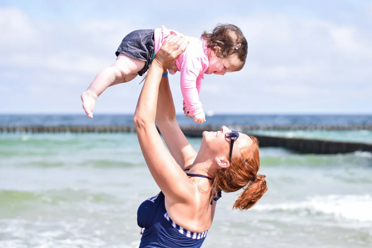 Radosna mama podrzuca dziecko nad morzem, ciesząc się wspólnym czasem i świeżym, jodowym powietrzem.