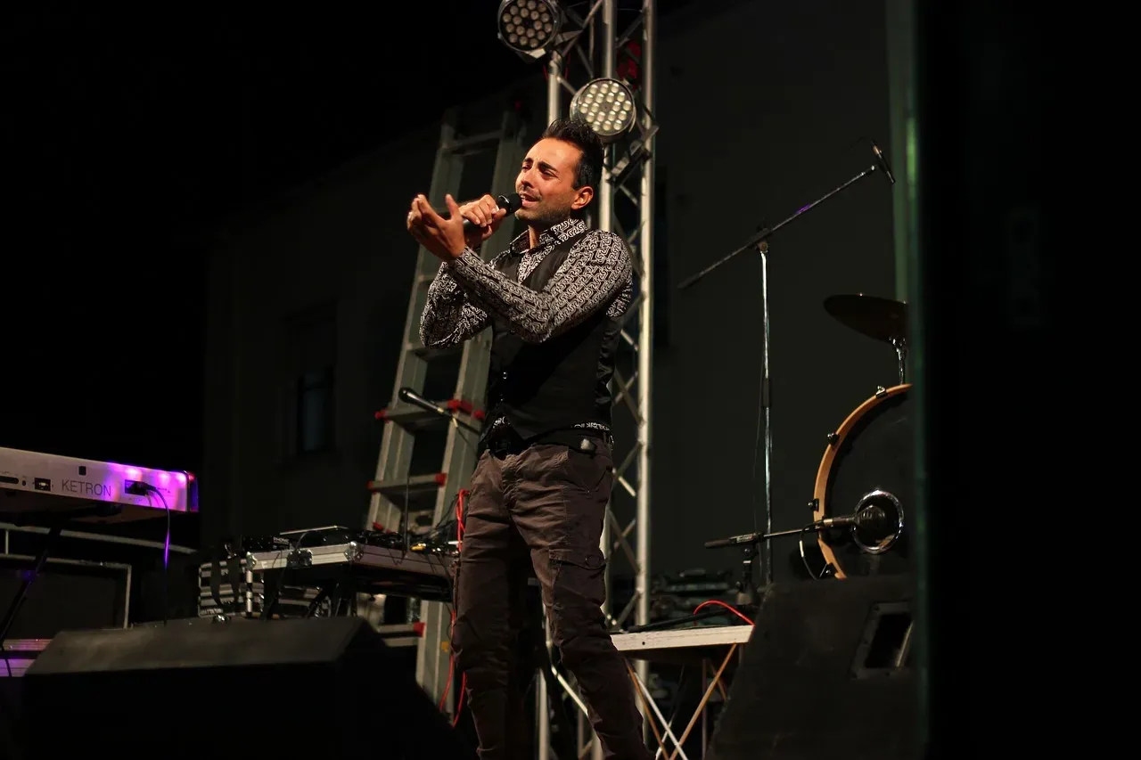 Wokalista na scenie, z mikrofonem w dłoni, w tle drabina i sprzęt muzyczny. Zastanawiasz się, ile trwa koncert?