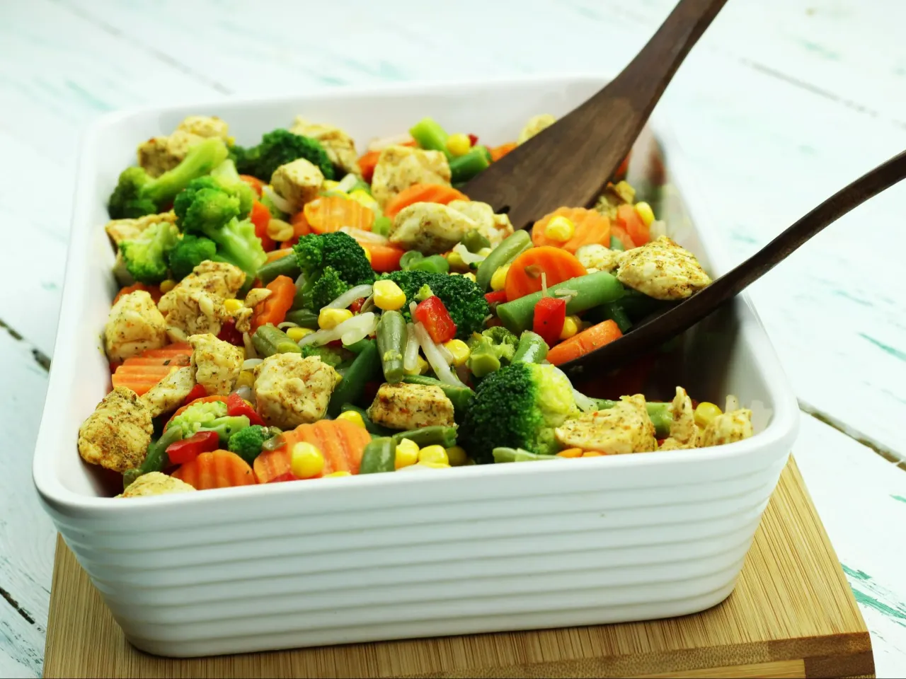 Kolorowa mieszanka warzyw z kurczakiem w białej misce, idealna jako przepis na zdrowy obiad. Drewniane łopatki mieszają danie.