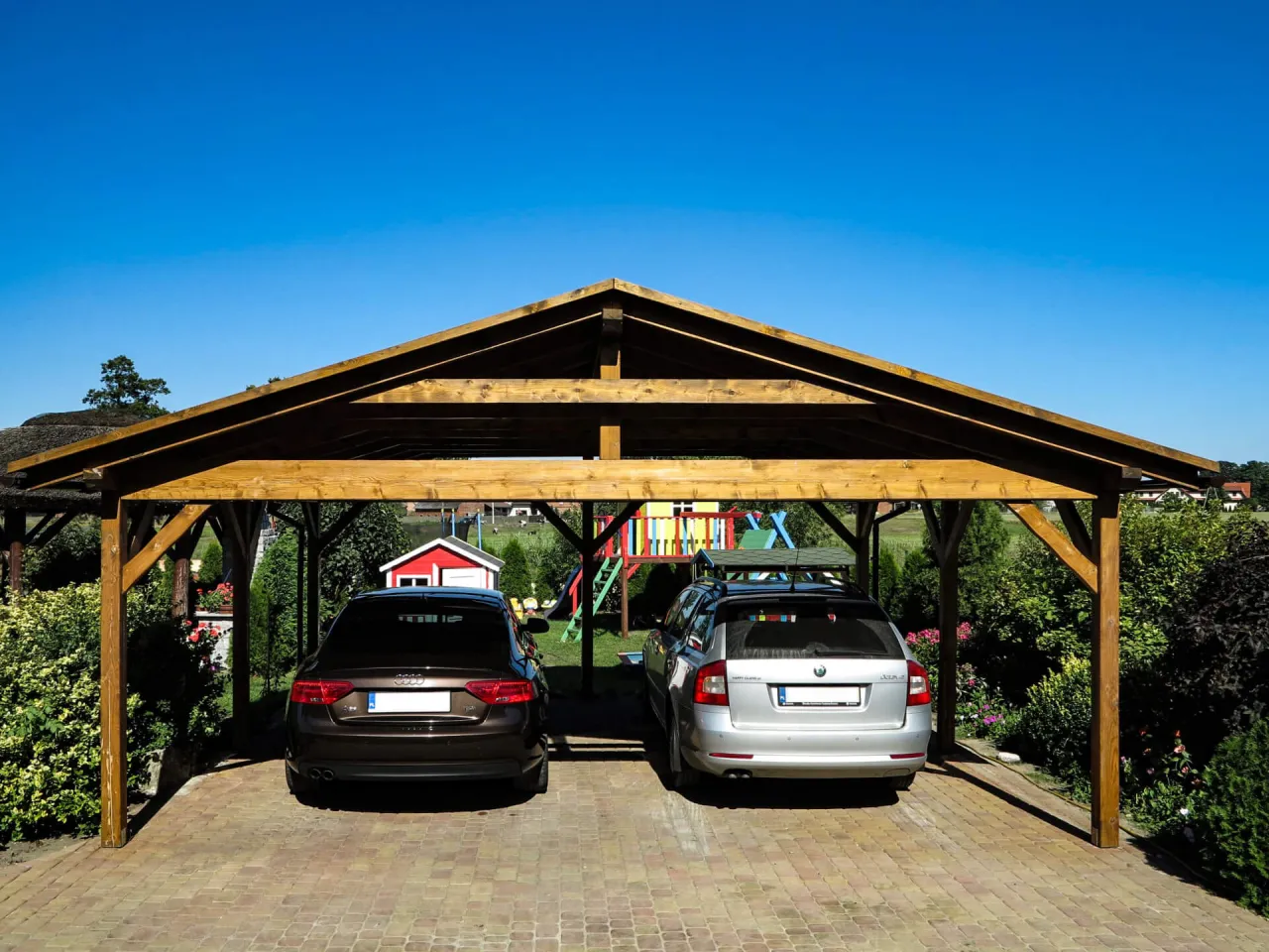 Dwa samochody pod solidnymi, drewnianymi wiatami garażowymi. W tle plac zabaw i ogród.