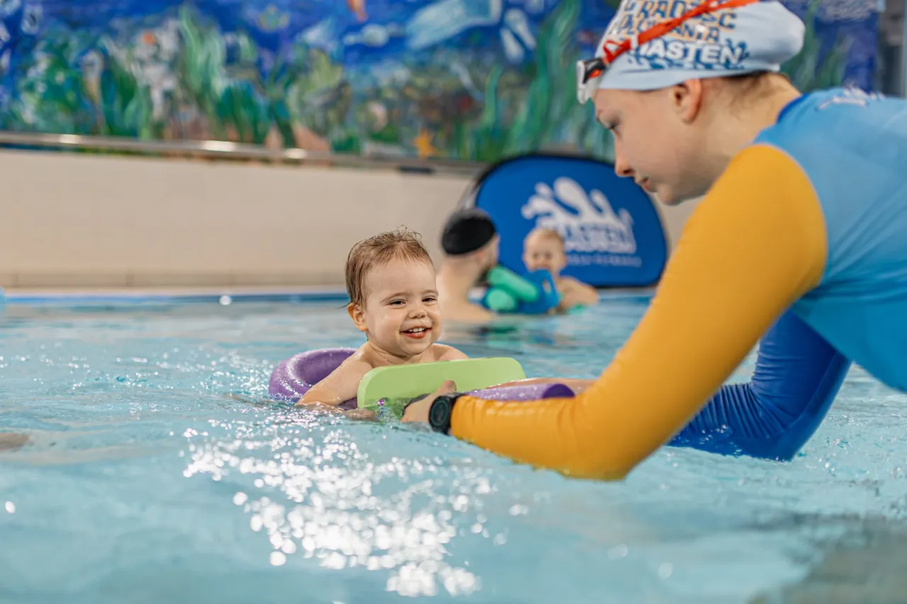 Uśmiechnięte dziecko w kółku do pływania, instruktorka podaje mu deskę. Dowiedz się, ile kosztuje indywidualna lekcja pływania dla malucha.