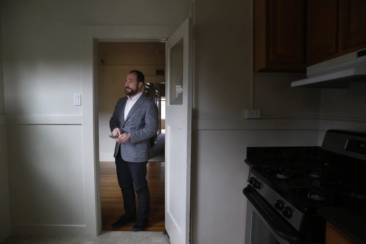 Mężczyzna w eleganckim garniturze stoi w progu kuchni, trzymając telefon. Zastanawia się, co to jest odstępne za mieszkanie.
