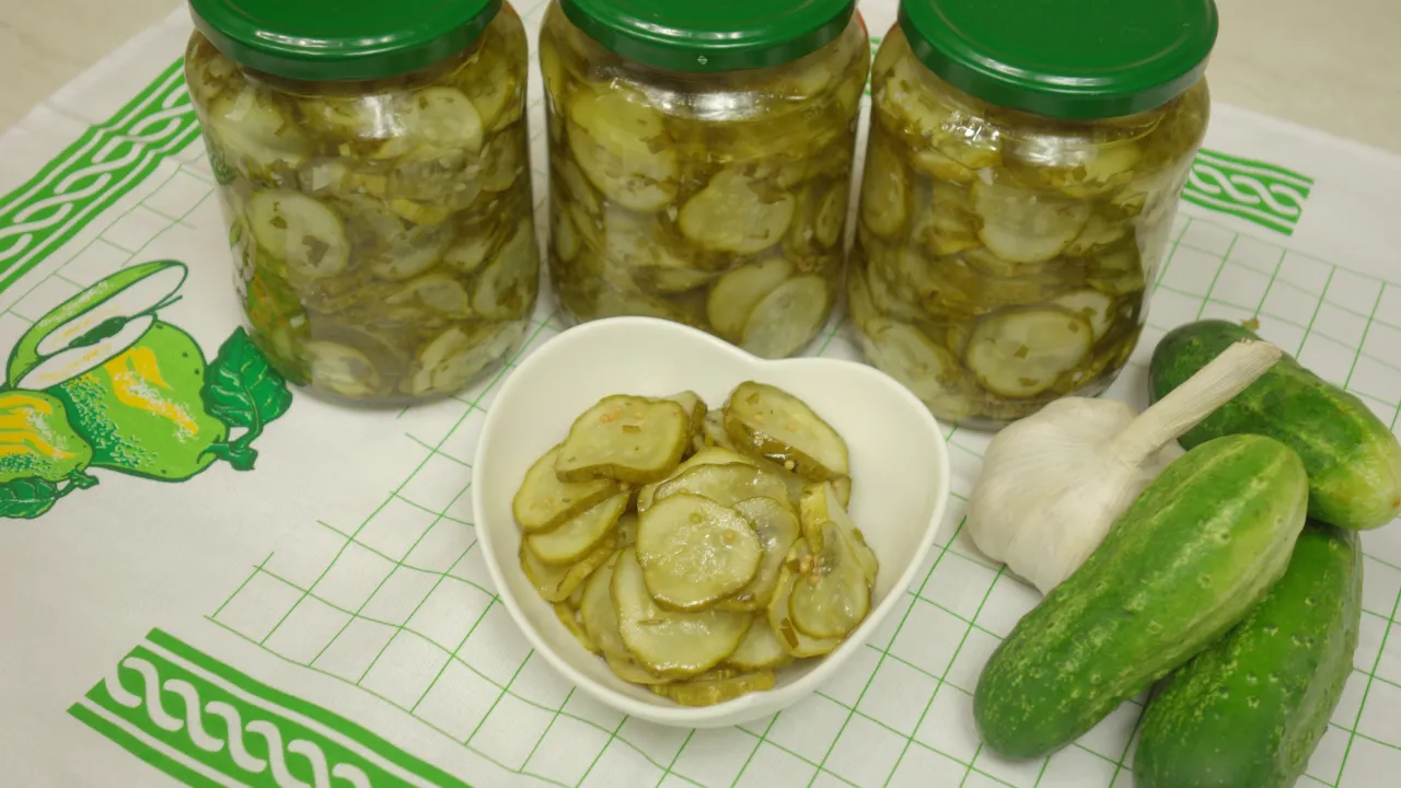 Słoiki z sałatką z ogórków, miseczka z plasterkami, świeże ogórki i czosnek. Idealne na zimę, gdy zastanawiasz się, ile pasteryzować sałatkę z ogórków.