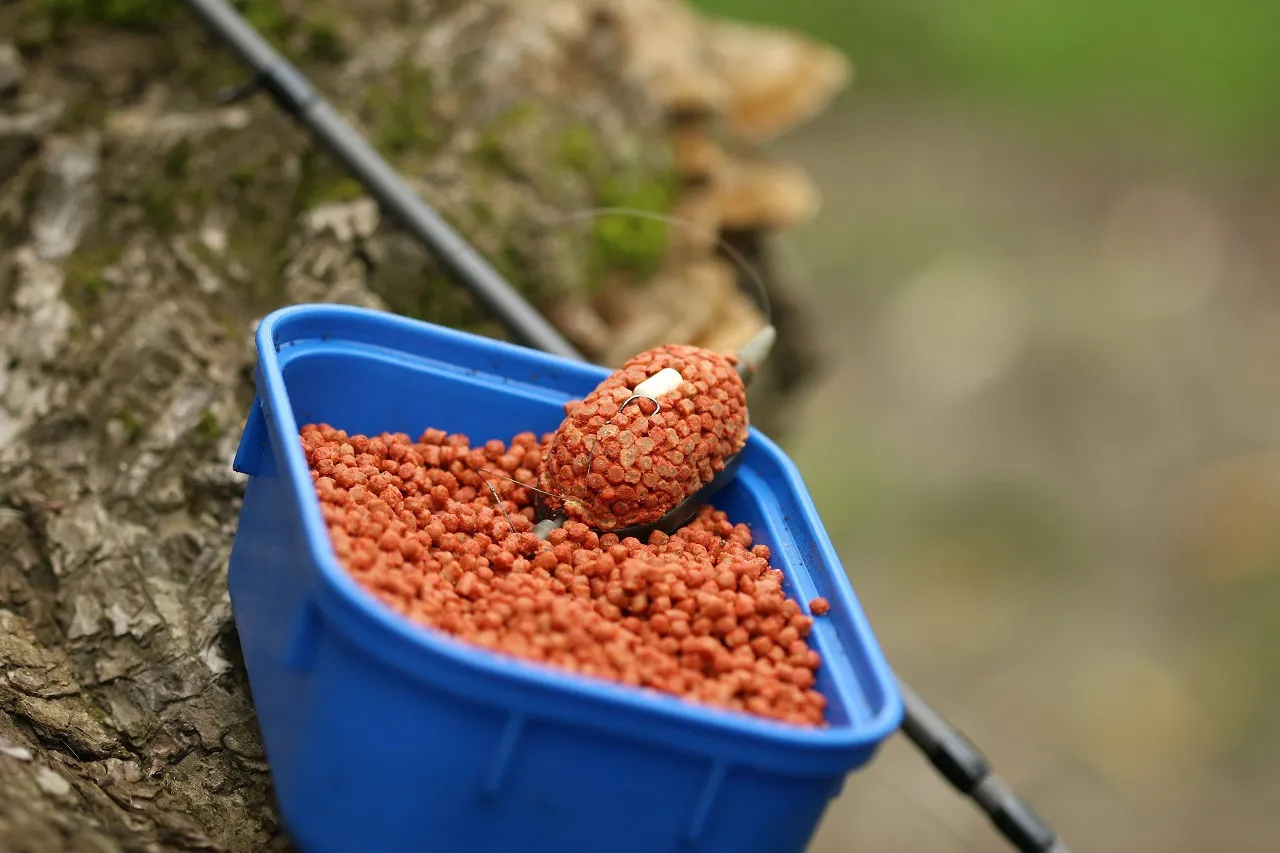 Niebieskie wiaderko z zanętą method feeder i na nim uformowana kulka z haczykiem. W tle wędka i drzewo.