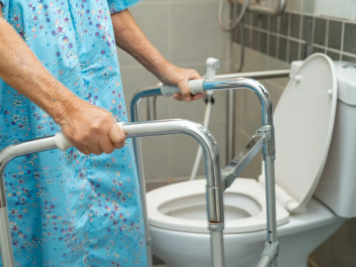 Starsza osoba w niebieskiej sukience z kwiatami, trzymająca się chodzika, obok toalety. Jak zmusić starszą osobę do mycia?