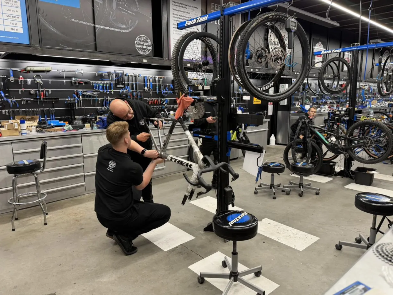 Mechanicy pracują przy rowerze, sprawdzając, ile kosztuje wymiana przerzutek w rowerze. W tle widać narzędzia i inne rowery.