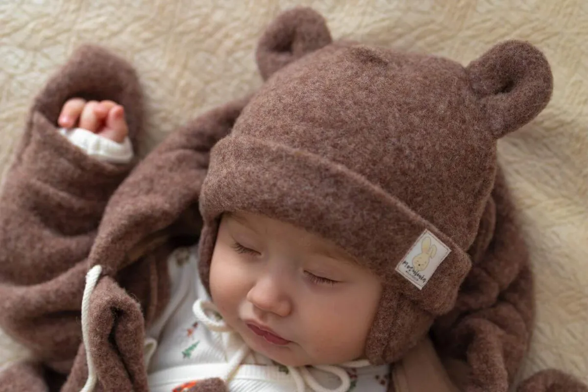 Śpiące dziecko w brązowej czapce merino dla dzieci z uszkami. Otulone w ciepły kombinezon.