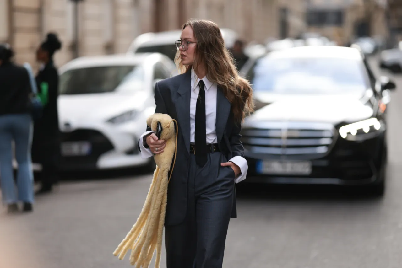 Kobieta w eleganckim garniturze i krawacie, z futrzaną torbą, prezentuje nowoczesny kobiecy dress code na tle miejskiego krajobrazu.