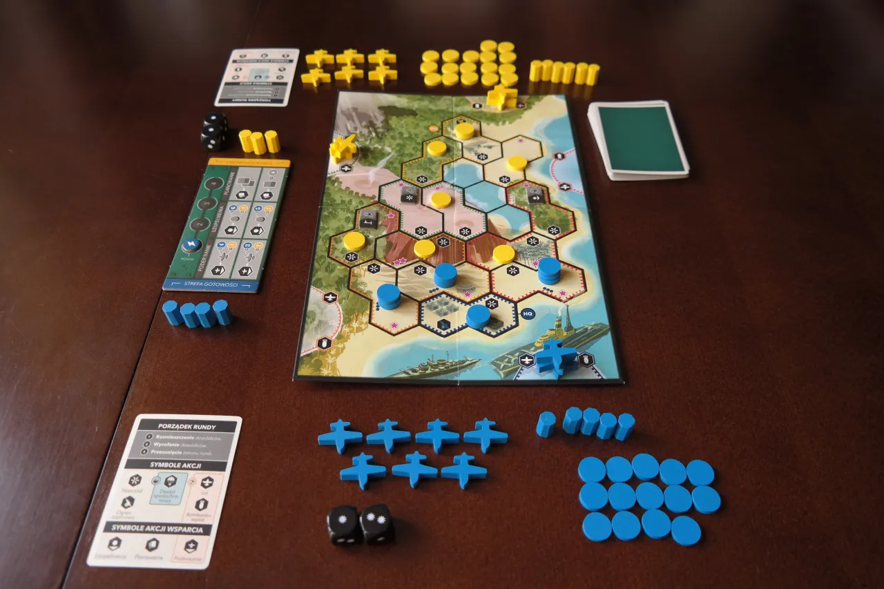 Na stole rozłożona jest plansza do gry, przypominająca mapę wyspy z morzem i g&oacute;rami. Widać żetony, kostki i karty. To gra planszowa, kt&oacute;ra wciąga graczy w strategiczne rozgrywki.