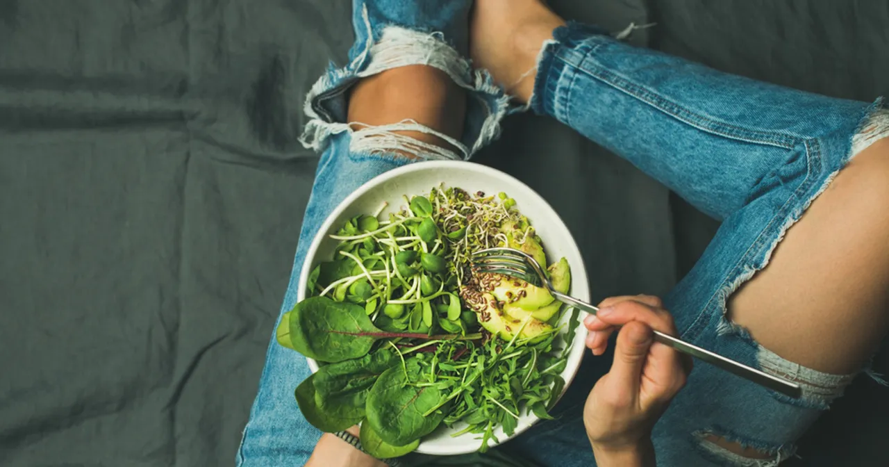 Osoba w podartych jeansach je sałatkę z awokado, kiełkami i zielonymi liśćmi. To przykład tego, na czym polega dieta wegańska.