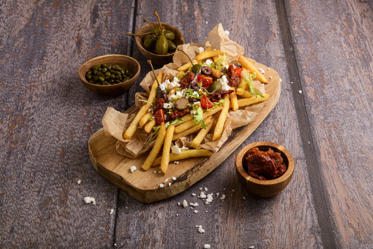 Złociste frytki z fetą, pomidorkami, oliwkami i pesto. Obok miseczki z kaparami i suszonymi pomidorami.