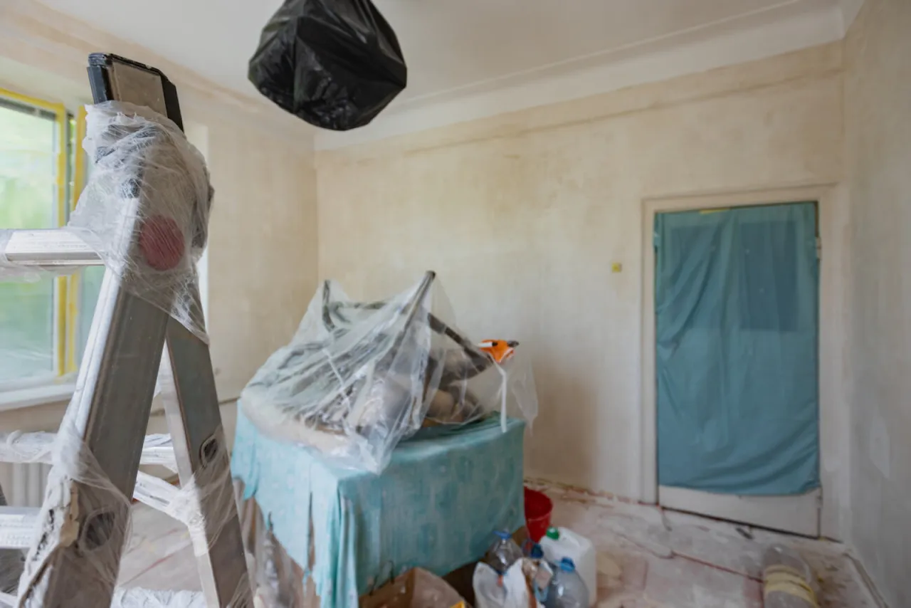 Pokój w trakcie remontu, przygotowany do malowania sufitu. Widać drabinę, meble przykryte folią i materiały budowlane.