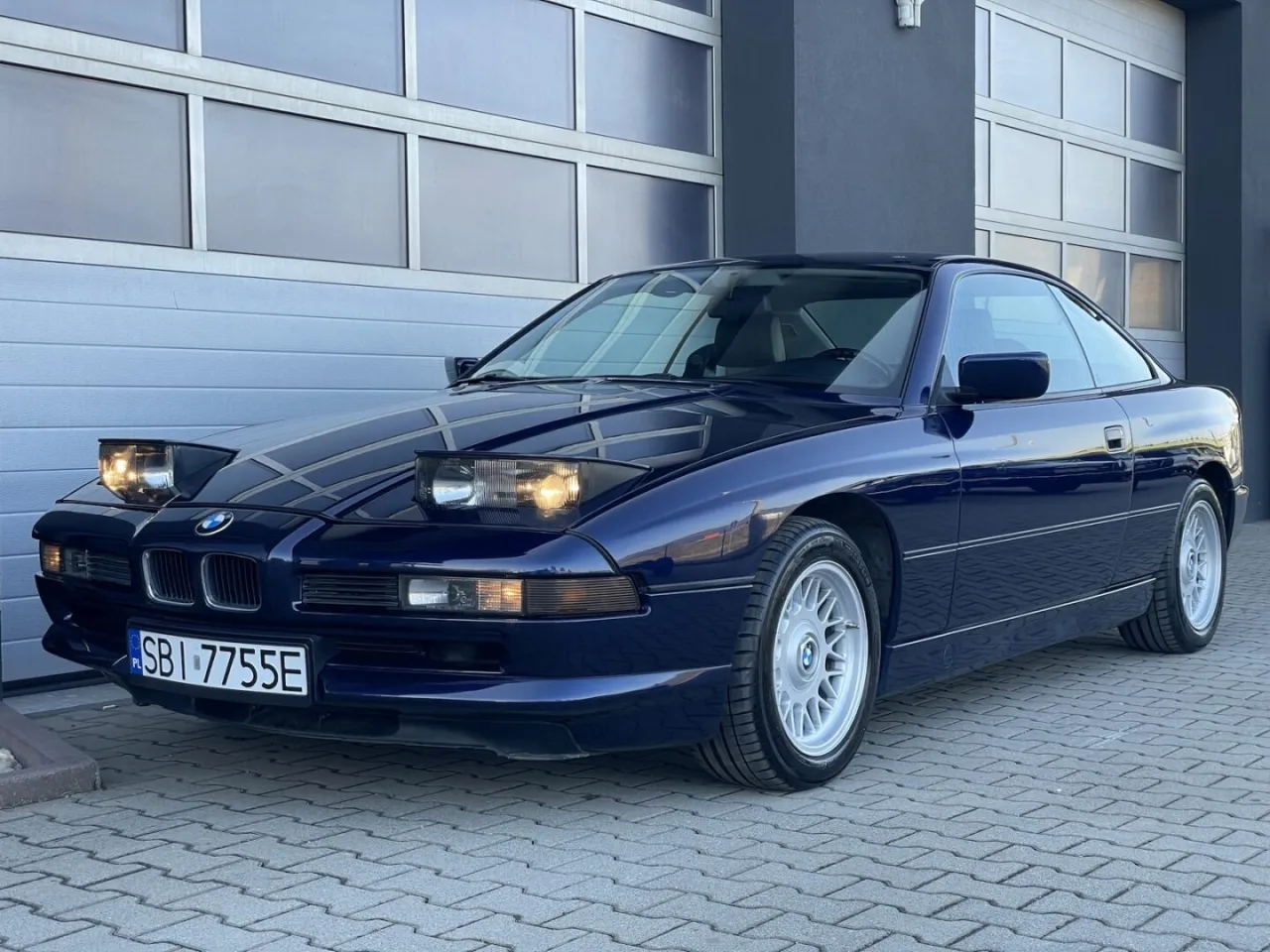 Granatowe BMW Serii 8 z silnikiem V12, z podniesionymi światłami, zaparkowane przed garażem.