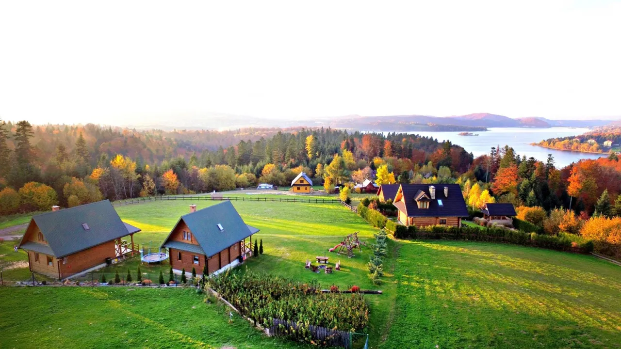 Jesienny krajobraz z domkami na zielonych wzg&oacute;rzach nad Soliną. Słońce przebija się przez mgłę, malując lasy w odcieniach złota i czerwieni.