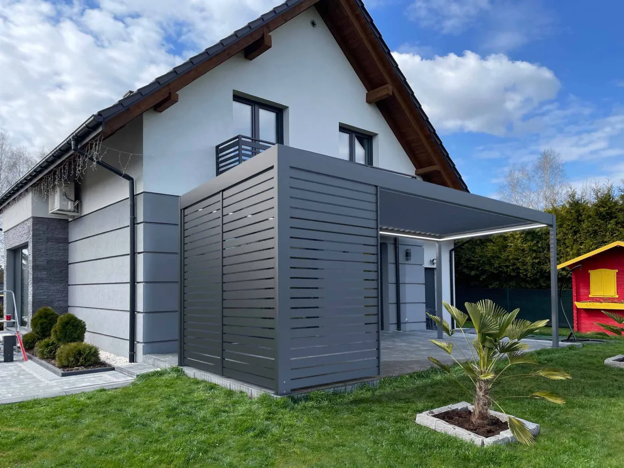 Nowoczesna pergola przy domu. Zastanawiasz się, ile kosztuje zrobienie pergoli? Ta imponująca konstrukcja z szarego aluminium z pewnością podnosi wartość nieruchomości.