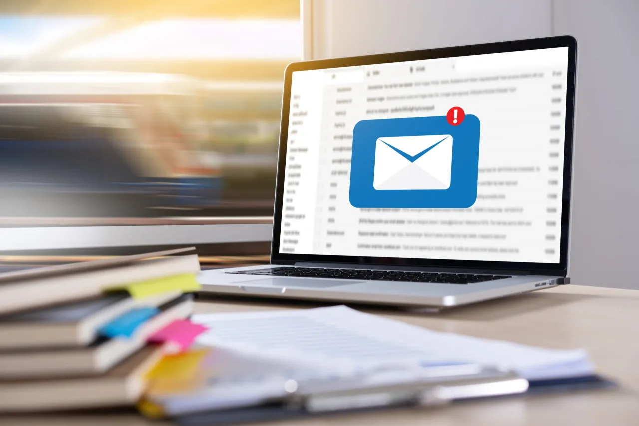 Laptop z ikoną koperty i wykrzyknikiem. Czy odpowiedź na pozew doręcza się pełnomocnikowi? Stos książek i dokumentów na biurku.