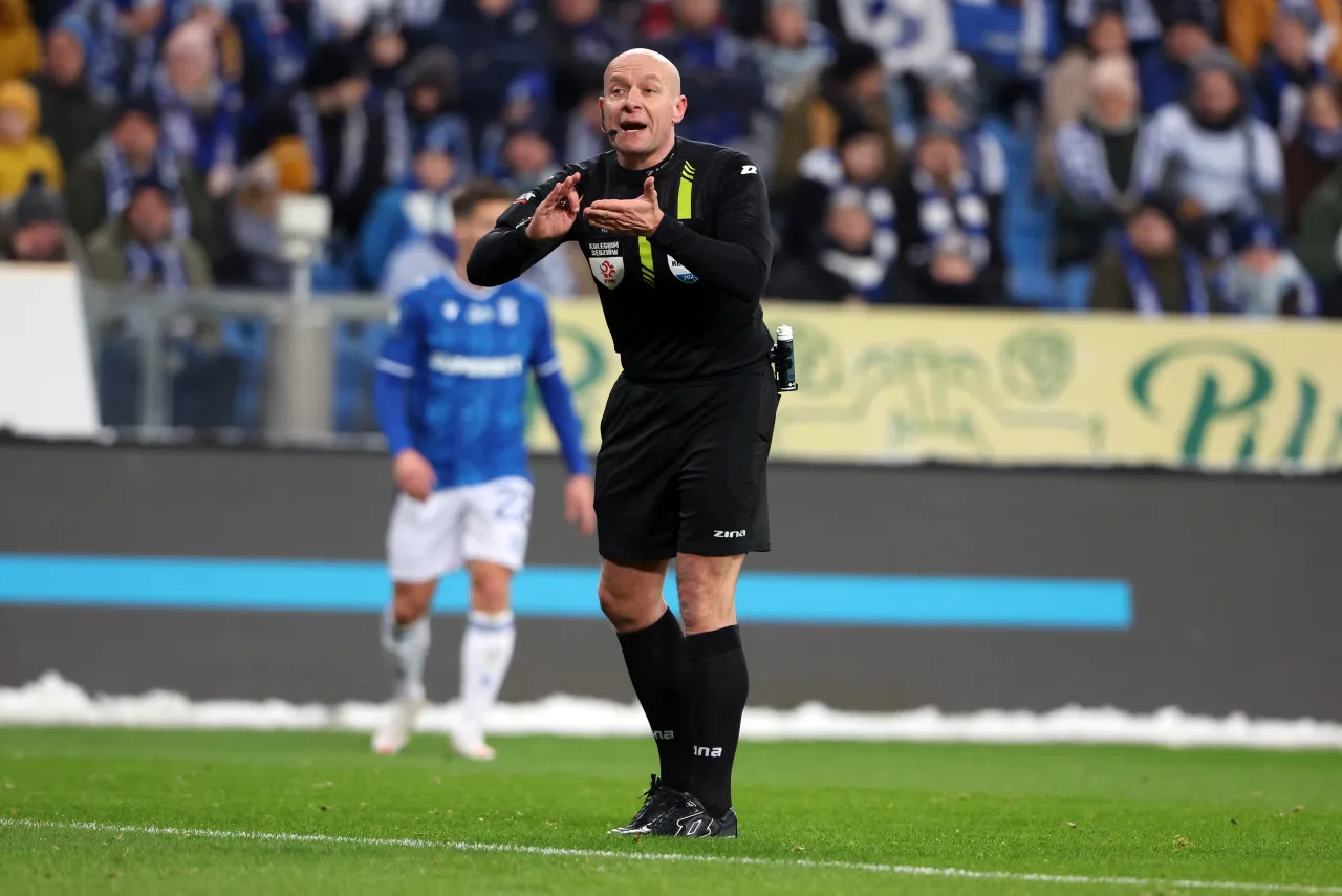 Sędzia w Ekstraklasie gestykuluje, być może zastanawiając się, ile zarabia. Na boisku widać też piłkarza w niebieskiej koszulce.