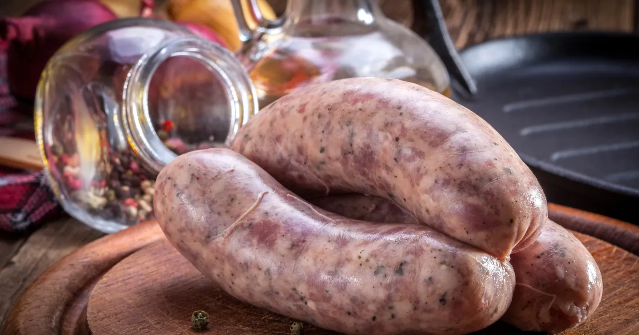 Surowa biała kiełbasa na desce, gotowa do parzenia. Obok słoik z przyprawami i patelnia.