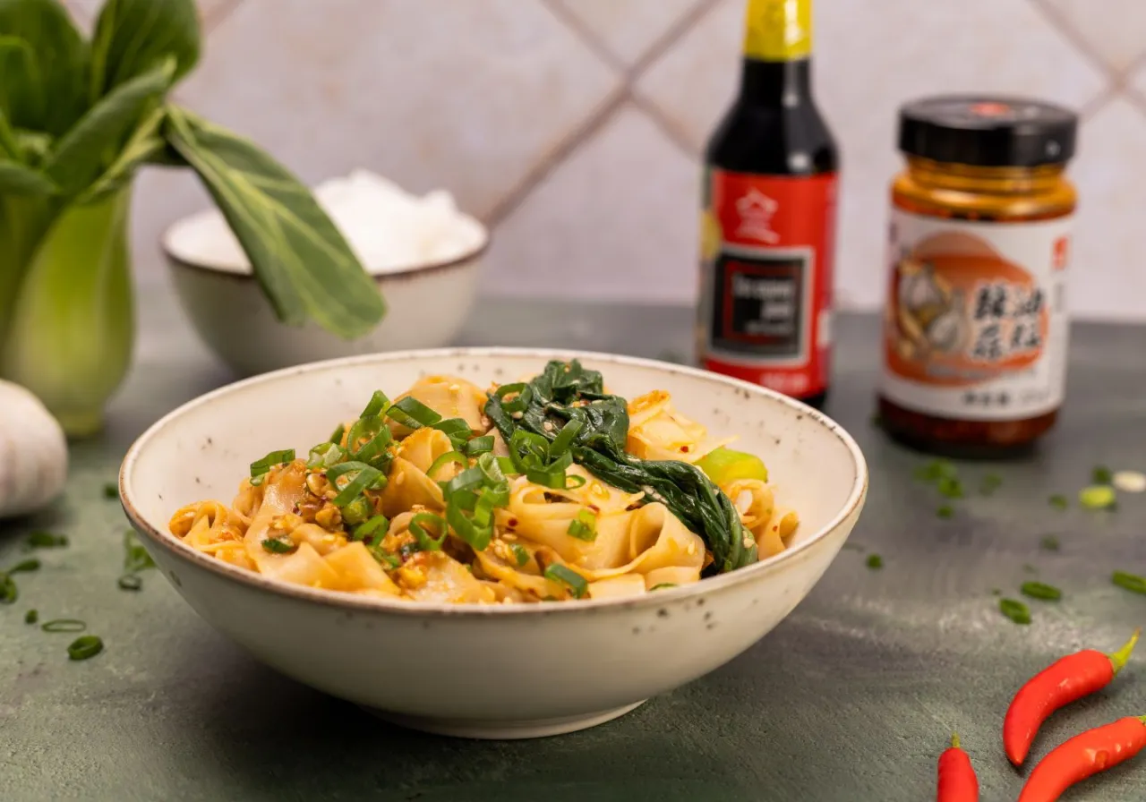 Miska szerokiego makaronu biang biang z zieloną cebulką i warzywami, obok sos sojowy, chili i czosnek.
