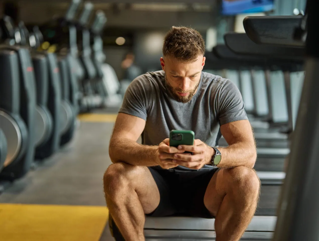 Mężczyzna w siłowni sprawdza telefon, zastanawiając się, ile trzeba ćwiczyć żeby schudnąć 5 kg.