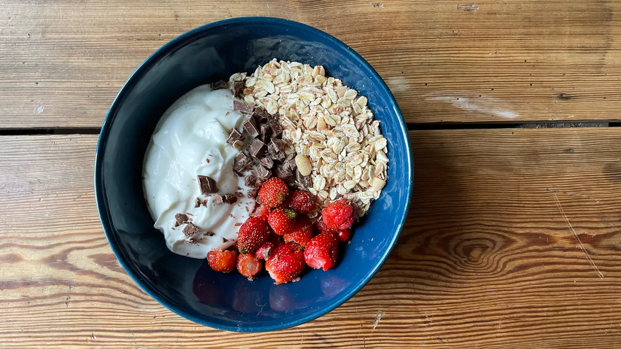 Jak zrobić dietetyczne musli: jogurt, płatki owsiane, truskawki i kawałki czekolady w niebieskiej misce na drewnianym stole.