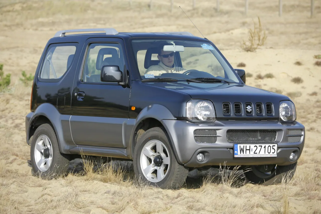 Czarny Suzuki Jimny na piaskowym podłożu. Kierowca w okularach. Pojemność baku tego modelu jest idealna na wyprawy.